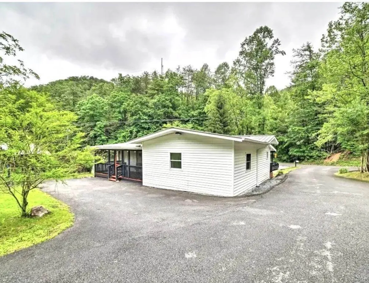 A cozy cabin nestles among vibrant greenery, with a spacious porch and driveway visible. Surrounded by nature, the house invites relaxation and hints at the comfort awaiting after outdoor adventures.