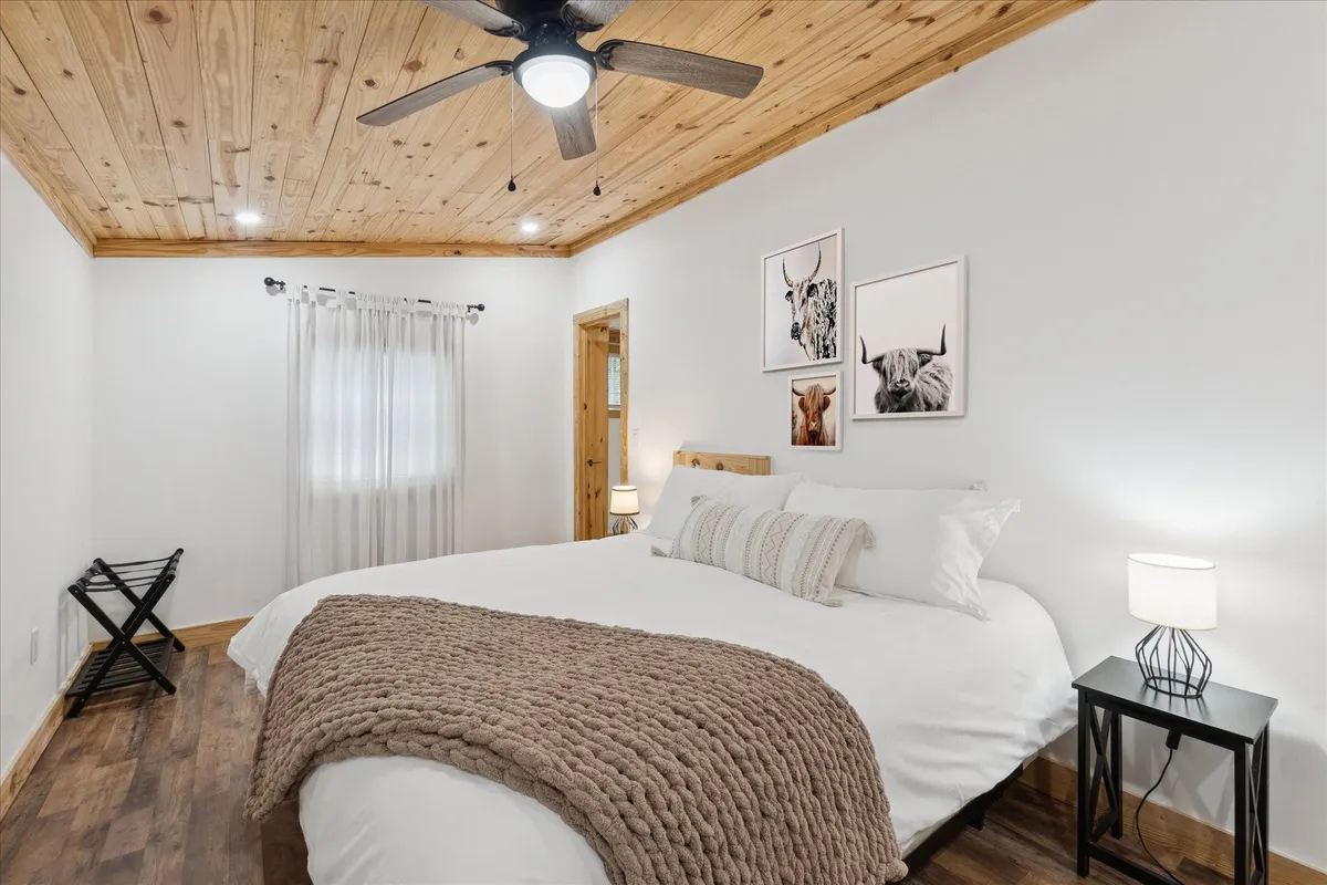 A serene bedroom with a comfortable bed, wooden ceiling, wall artwork, and nightstands on either side. Natural light softly illuminates the space, creating a warm, inviting atmosphere that blends rustic charm with modern touches.