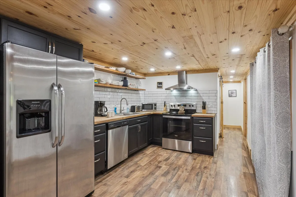 The image shows a cozy kitchen with stainless steel appliances, wooden ceilings, black cabinetry, and open shelving stocked with essentials. The layout feels functional yet inviting, perfect for preparing group meals or holiday feasts.