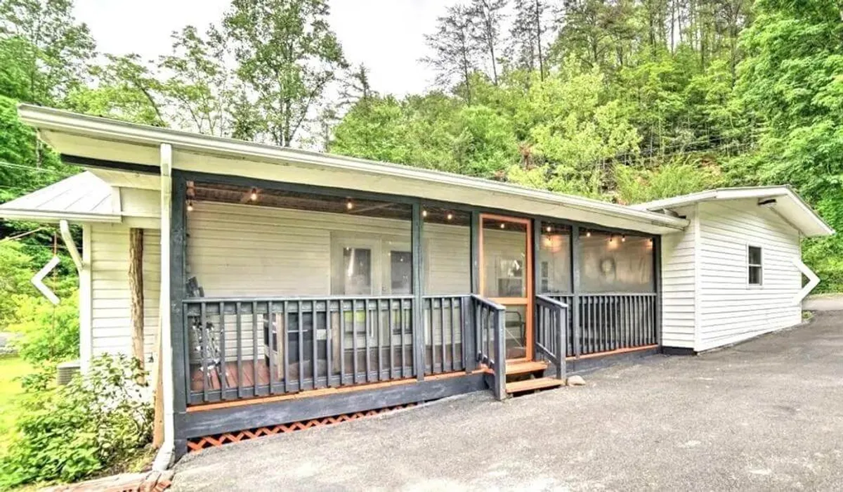A spacious, screened-in porch is framed by wooden railings and a welcoming front door. The porch is perfect for gathering friends to enjoy nature, bask in the fresh air, or savor coffee at sunrise or drinks at dusk—setting the stage for memorable group moments.
