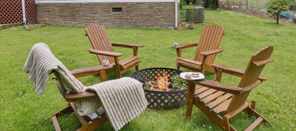 A stone fire pit surrounded by cozy Adirondack chairs draped with blankets, with the seating arranged in a circle for easy conversation. Twinkling string lights above add warmth and charm, while the darkening sky hints at a starlit evening. The setting encourages guests to gather, share stories, and toast marshmallows together.