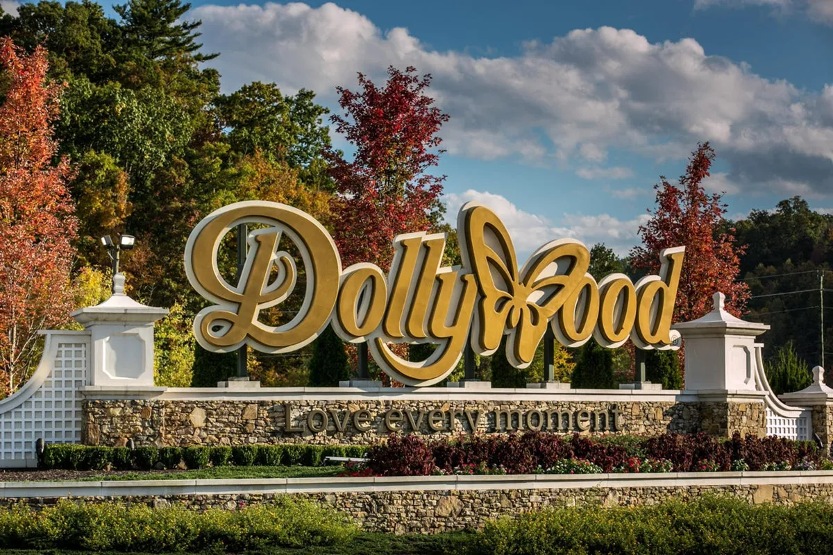 This image captures the iconic Dollywood sign surrounded by vibrant, colorful trees and a stone wall at the park's entrance. The lively atmosphere suggests the anticipation and joy of arriving at a major family-friendly attraction.