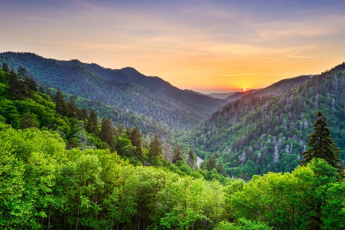 A breathtaking view of a Smoky Mountain range at sunset, with a winding river cutting through the valley below. The sky is painted with warm hues, emphasizing the vastness and tranquility of the natural surroundings.