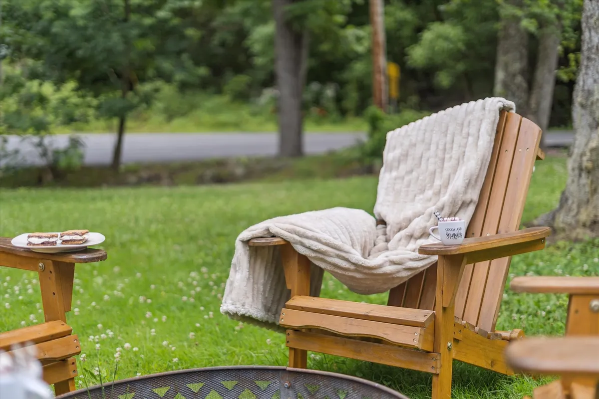 A wooden chair, draped with a soft blanket, faces a glowing fire pit. On the table beside it sits a steaming coffee mug and a plate of snacks, suggesting a tranquil morning or evening by the fire. The natural backdrop and thoughtful touches evoke relaxation and warmth.